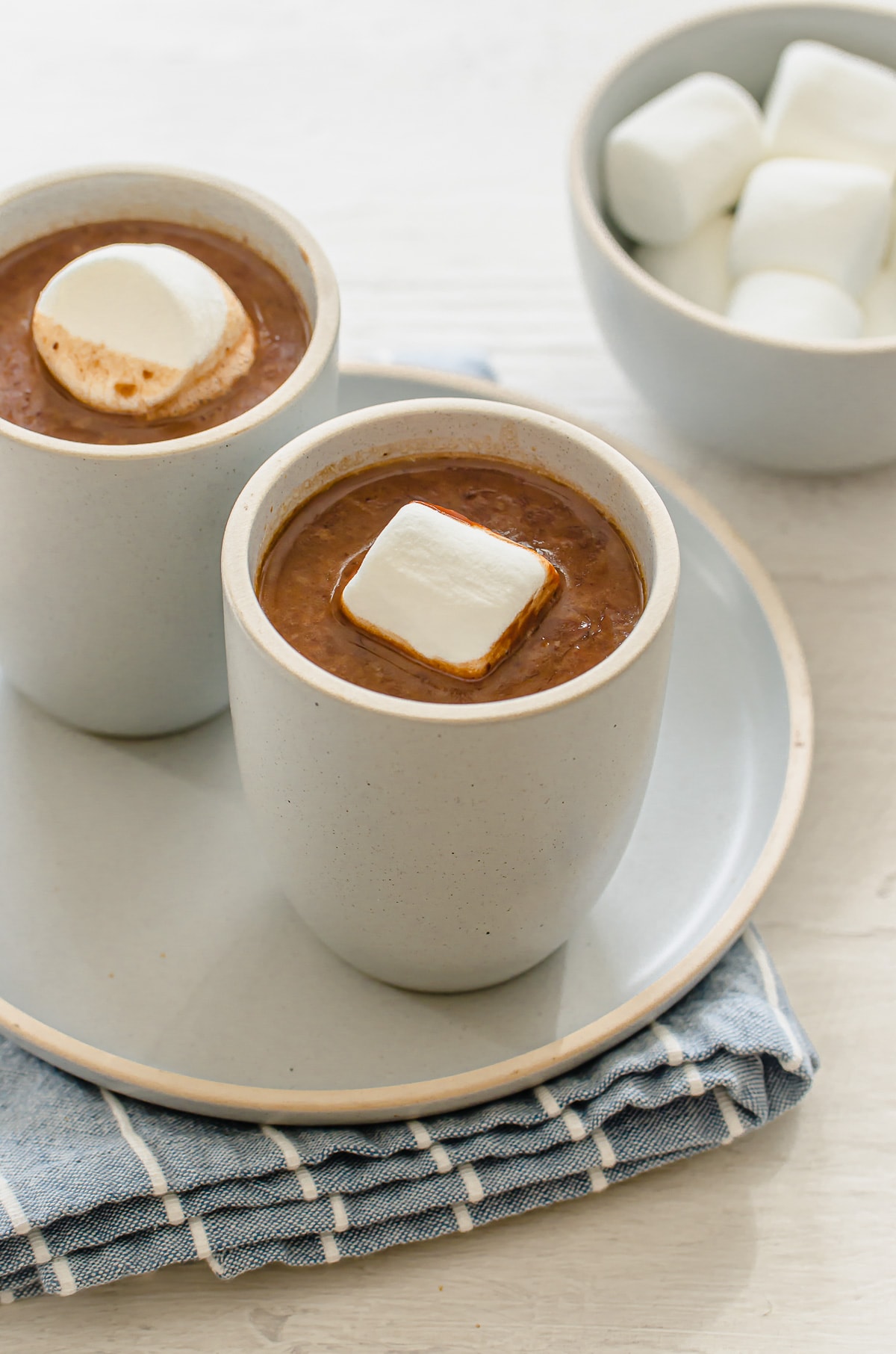 A side shot of two blue ceramic cups of hot chocolate with large marshmallows on top with a bowl of marshmallows in the background.