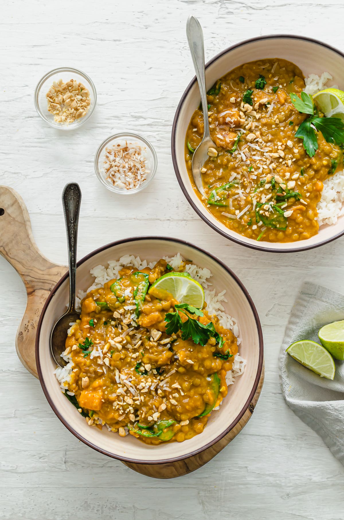 An overhead shot of bowls of lentil curry with toppings of toasted coconut, chopped peanuts, and fresh lime wedges on the side.