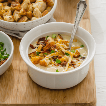 A white bowl of potato leek soup topped with croutons, bacon, cheese, and scallions.